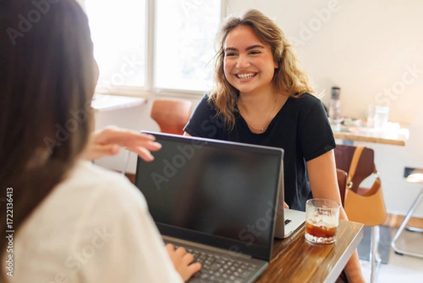 Fototapeta Two young women engage in lively conversation while working in a cozy cafe