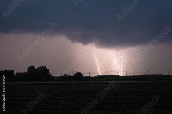 Fototapeta Dramatic lightning storm over open field at night with silhouetted trees and fence