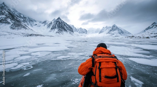 Fototapeta Individual in vibrant orange jacket is navigating icy terrain surrounded by towering snow-capped mountains and a serene atmosphere