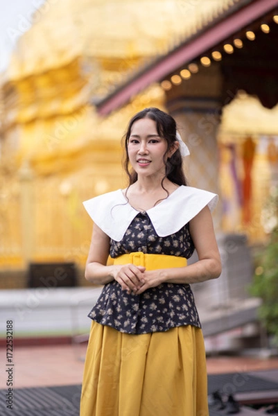 Fototapeta Asian woman in a retro-inspired outfit with a yellow belt poses with a gentle smile in front of a golden Buddhist temple.