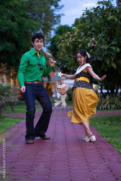 Fototapeta A young couple dressed in colorful retro-style outfits dancing together on a red brick walkway in a garden.