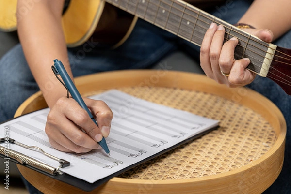 Fototapeta A creative young woman is enjoying her hobby, skillfully playing a guitar while concentrating on writing musical notes in her notebook. The scene captures the essence of music composition and artistic