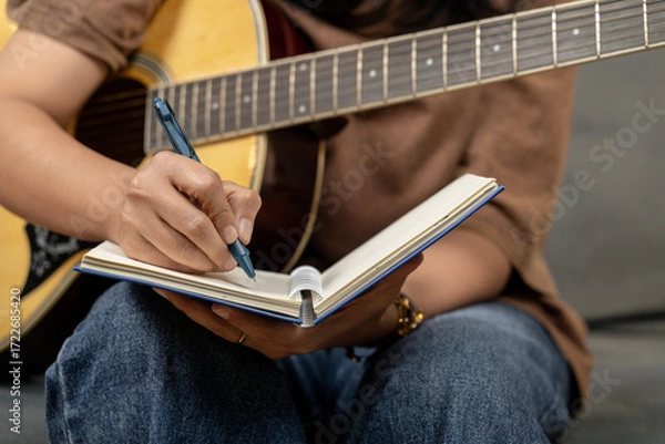 Fototapeta A creative young woman is enjoying her hobby, skillfully playing a guitar while concentrating on writing musical notes in her notebook. The scene captures the essence of music composition and artistic