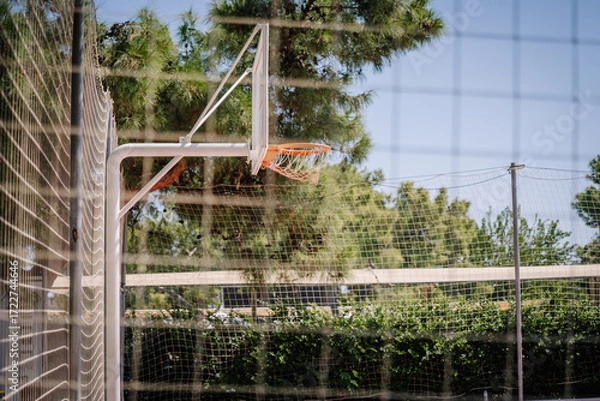 Fototapeta Close-up of a basketball hoop on an outdoor court surrounded by netting and green trees, under a bright sunny sky in a park or recreational area.