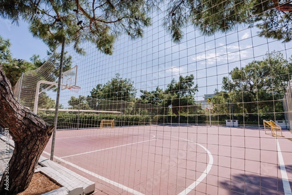 Fototapeta Outdoor basketball and soccer court surrounded by a safety net, trees, and greenery, under a clear blue sky on a sunny day in a public park.