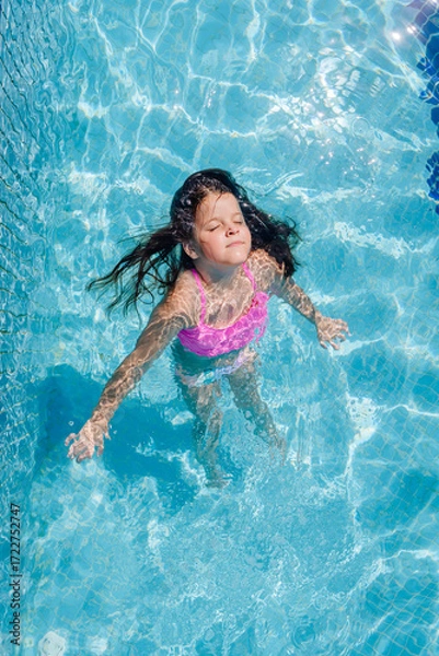 Fototapeta Young girl in a pink swimsuit floating peacefully in a swimming pool with closed eyes, arms extended, enjoying sunlight and summer fun in clear blue water.