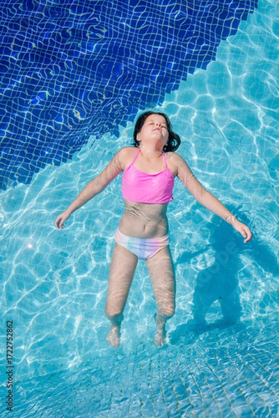 Fototapeta Little girl floating in a swimming pool with eyes closed, arms outstretched, and peaceful expression, enjoying a sunny summer day in colorful swimwear.
