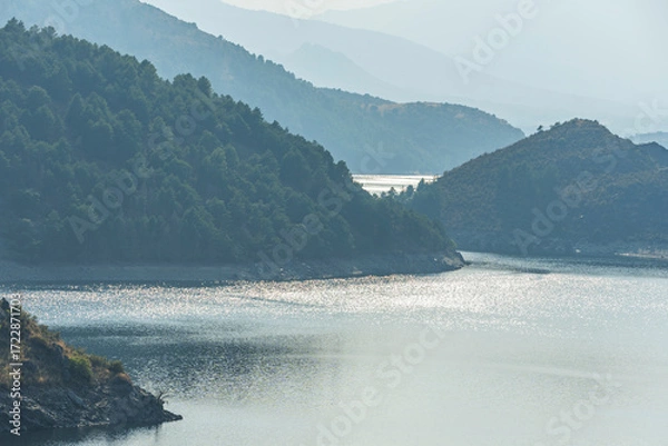 Fototapeta Panoramic view of the Atazar Reservoir at sunset, with blue waters surrounded by mountains and orange skies reflecting on the surface