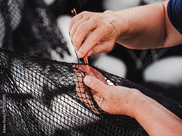 Fototapeta Skilled hands repair fishing net in coastal workshop during bright afternoon light