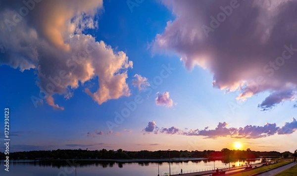 Fototapeta Pink and orange clouds float magically over the river at sunset. Panorama of calm evening fluffy sky near the embankment. Walk before bed, enjoying cloud nature evening. Yellow sun reflection water.