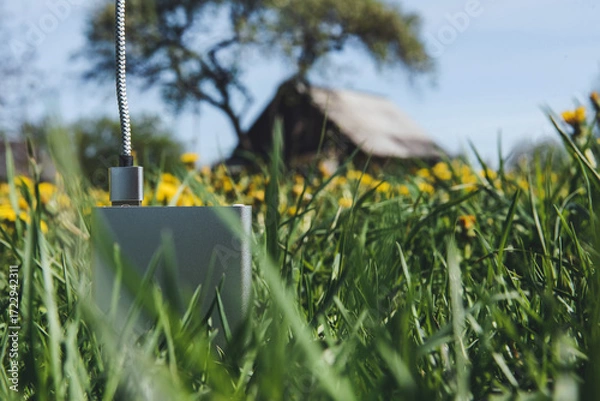 Obraz Silver power bank in green grass with a blurry rustic house background.