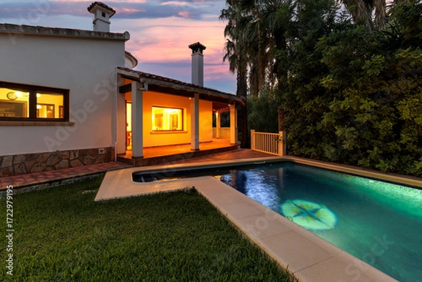 Obraz Evening view of a Mediterranean-style villa across an illuminated pool and green lawn.