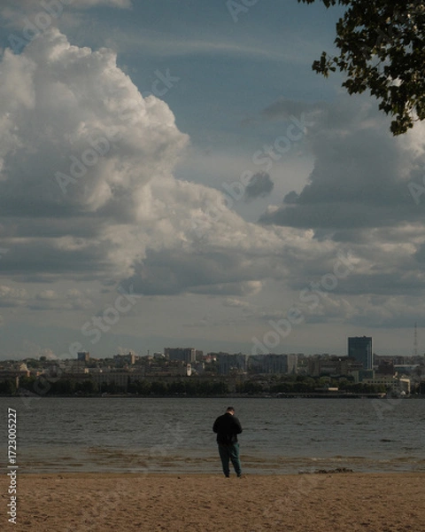 Fototapeta A view of a cityscape across a river or body of water on an overcast day. In the foreground, on a sandy shore, stands a lone person. Thick clouds in the sky create a pensive atmosphere.