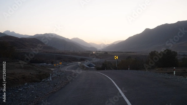 Fototapeta The winding road goes somewhere into the distance, mountain peaks are visible in the distance, twilight, evening panorama of the mountain crevice and the road ahead