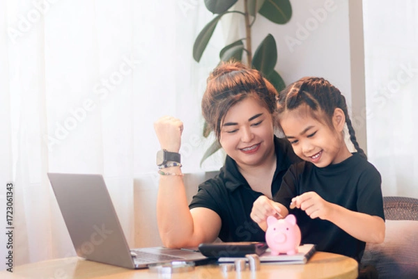 Obraz Fun savings lesson between mother and daughter at home with piggy bank and laptop during the day