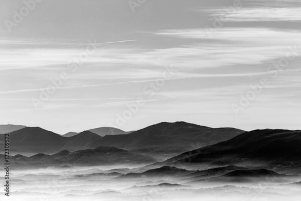 Fototapeta A view from above of a valley filled by a sea of fog, with various layers of emerging hills and mountains