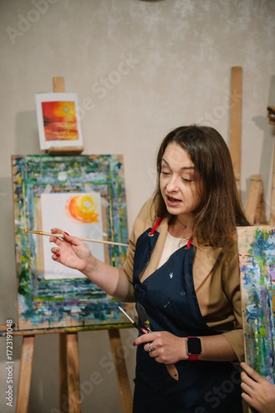 Fototapeta Teacher learn children to draw pictures in an art lesson. Painting in workshop. Artist explains how to mix and combine colors. Woman drawing brush on easel. Education, learning concept. Closeup.
