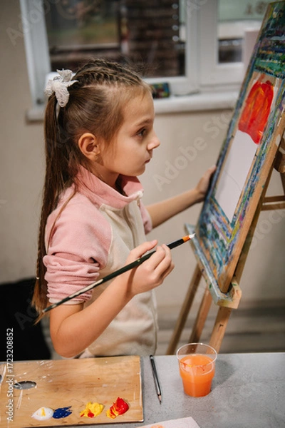 Fototapeta Girl enjoys painting in workshop. Art lesson. Education and learning concept. Child draw a picture in art class. School kid drawing with brush landscapes on easel. Mix and combine colors.