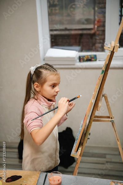 Fototapeta Child draw a picture in art class. Girl enjoys painting in a workshop. Art lesson. Education and learning concept. School kid drawing with brush landscapes on easel. Mix and combine colors.