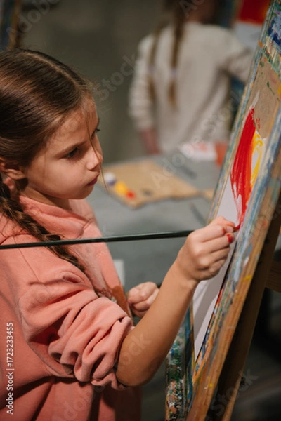 Fototapeta Girl enjoys painting in workshop. Art lesson. Education and learning concept. Child draw a picture in art class. School kid drawing with brush landscapes on easel. Mix and combine colors.