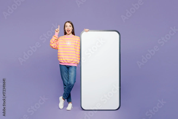 Fototapeta Full body young amazed woman wears yellow sweatshirt casual clothes big huge blank screen mobile cell phone smartphone with area point finger up isolated on plain purple background. Lifestyle concept.