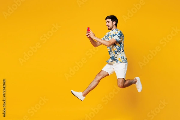 Fototapeta Full body side view young man he wearing blue beach t-shirt casual clothes jump high hold in hand use mobile cell phone isolated on plain yellow orange background studio portrait. Lifestyle concept.