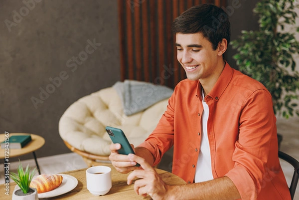 Fototapeta Side view young man he wear orange casual clothes use cell phone sitting alone at table in coffee shop cafe relax rest in restaurant during free time indoors. Freelance mobile office business concept.