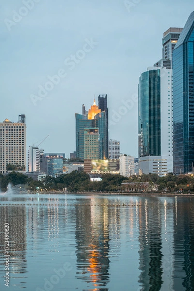 Fototapeta evening panorama of Bangkok city skyline with skyscrapers and Benchakitti park lake, vibrant cityscape view with reflections over pond water at dusk