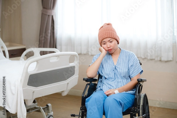 Fototapeta A female patient wearing a beanie and hospital gown sits in a wheelchair in a hospital room beside a bed, looking thoughtful and somewhat somber. The scene conveys rest,
