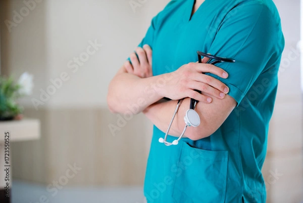 Fototapeta Close-up of unrecognizable doctor holding stethoscope while standing with arms crossed at medical clinic.