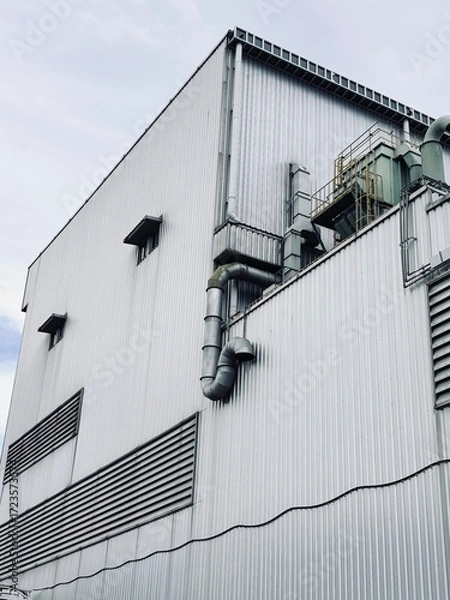 Fototapeta A monochrome, low-angle shot of an industrial building, highlighting the corrugated metal siding and a complex network of air ducts and pipes against a stark, empty sky.