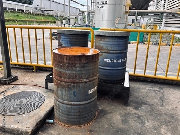 Fototapeta Three weathered industrial drums, two blue and one rusty, are placed in a service yard. The scene highlights themes of heavy industry, waste management, and manufacturing, with a factory building 