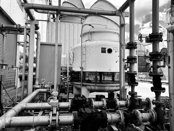 Fototapeta A black and white close-up of a complex industrial cooling system. The image highlights a dense network of pipes, valves, and a large cooling tower, capturing the intricate infrastructure of a factory