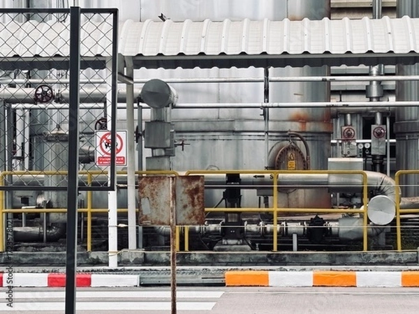 Fototapeta A detailed shot of a complex industrial facility, featuring a network of pipes, tanks, and machinery set against a gray metal building, conveying a sense of manufacturing and production.