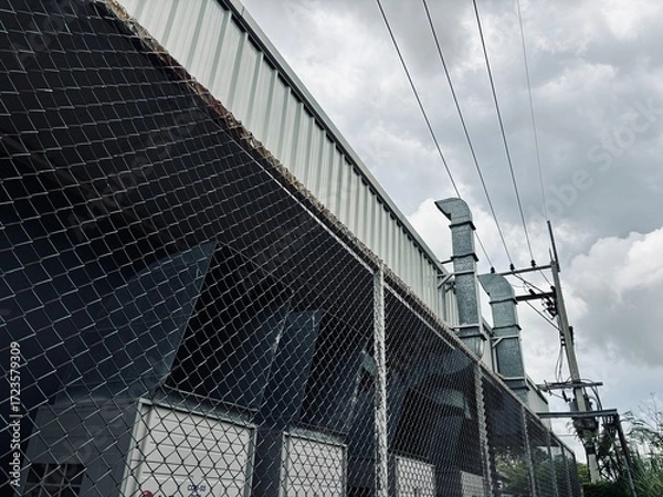 Fototapeta A low-angle shot of a corrugated metal warehouse or factory building with a rusty chain-link fence in the foreground, under a dramatic, cloudy sky.