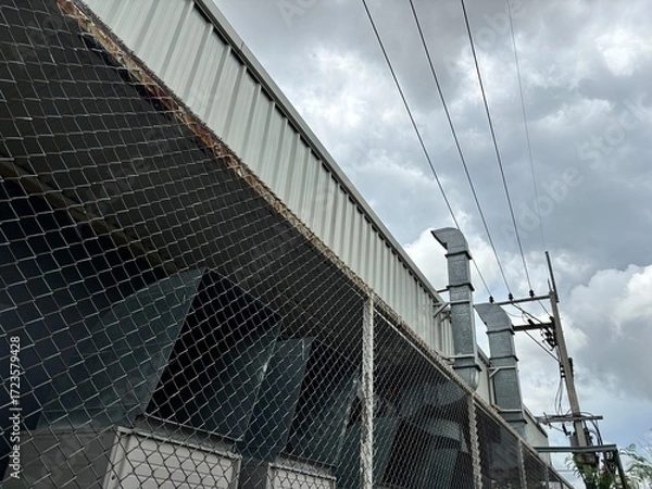 Fototapeta A low-angle shot of a corrugated metal warehouse or factory building with a rusty chain-link fence in the foreground, under a dramatic, cloudy sky.