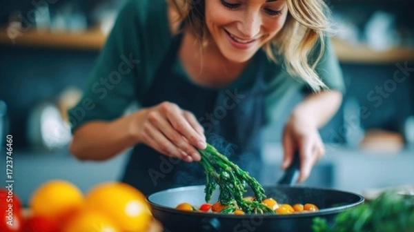 Fototapeta A woman is cooking in the kitchen, adding asparagus to a pan with other vegetables, smiling with joy.
