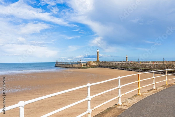Fototapeta A pier with lighthouses stretches out to sea with a beach and railings in the foreground.