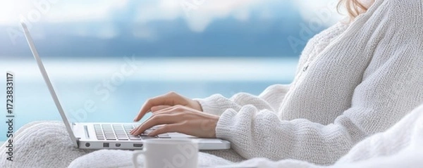 Fototapeta A cozy scene of a person typing on a laptop while wrapped in a soft sweater, with a serene background of water and gentle colors.