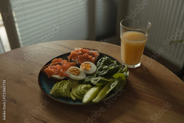 Fototapeta Nutritious breakfast plate with salmon sandwiches, boiled egg, avocado slices, cucumber, and fresh spinach served with glass of orange juice. Concept of healthy lifestyle, diet food, and balanced