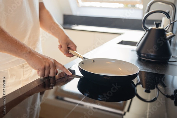 Fototapeta Close-up of man in white clothes putting frying pan on electric stove in modern kitchen. Concept of cooking, daily routine, and home lifestyle. Symbol of preparation, comfort, minimalistic interior