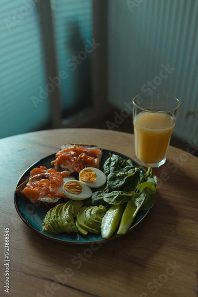 Fototapeta Nutritious breakfast plate with salmon sandwiches, boiled egg, avocado slices, cucumber, and fresh spinach served with glass of orange juice. Concept of healthy lifestyle, diet food, and balanced