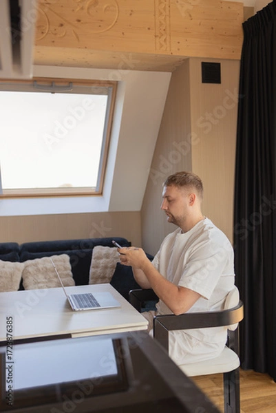 Fototapeta Young man sitting at desk with laptop and smartphone in modern home office. Concept of remote work, online communication, fintech, digital banking, e-commerce, productivity and technology lifestyle