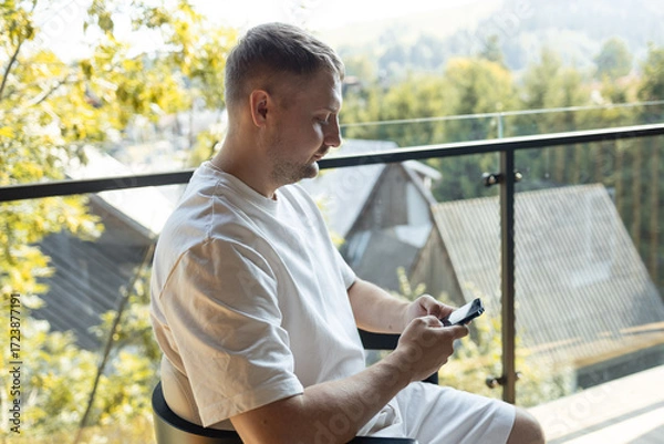 Fototapeta Casual man sitting on balcony in white clothes and looking at smartphone with scenic view of nature and houses in background. Concept of modern lifestyle, technology, and relaxation. Symbol of digital