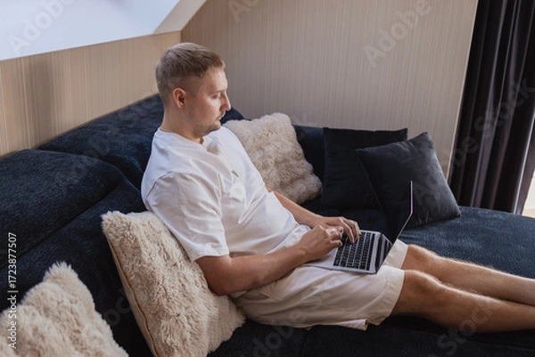 Fototapeta Young man sitting on sofa with laptop and smartphone, working remotely or managing online tasks. Concept of freelancing, digital lifestyle, mobile technology, e-banking, online shopping, communication