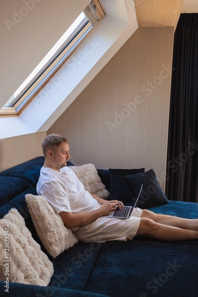 Fototapeta Young man sitting on sofa with laptop and smartphone, working remotely or managing online tasks. Concept of freelancing, digital lifestyle, mobile technology, e-banking, online shopping, communication