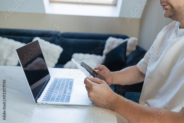 Fototapeta Young man sitting at home, using laptop, smartphone and credit card for secure online payment. Concept of digital banking, remote work, financial planning and modern technology in everyday life.