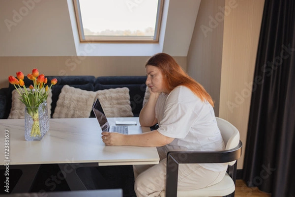 Fototapeta Woman sitting at desk with laptop, leaning head on hand with tired and bored expression. Concept of remote work challenges, lack of motivation, burnout, digital fatigue, procrastination, financial