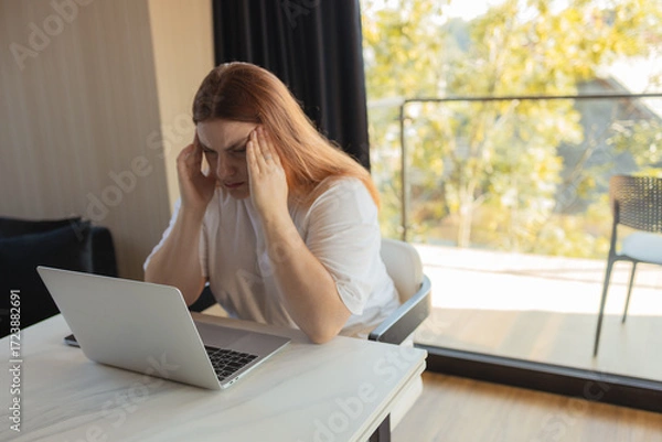 Fototapeta Exhausted woman sitting at desk with laptop, touching her forehead in stress. Concept of burnout, digital fatigue, remote work challenges, mental health, financial worries and technology dependence