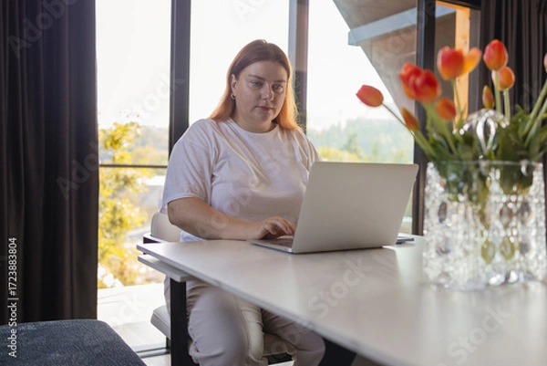 Fototapeta Young woman sitting at desk with laptop in cozy home interior, working remotely or studying online. Concept of freelancing, digital lifestyle, remote work, e-learning, communication, modern technology